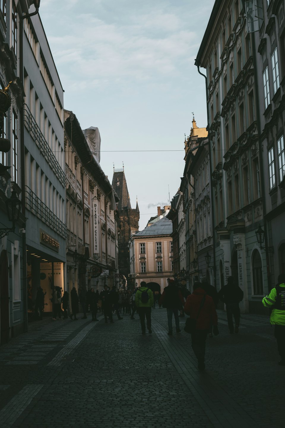 Pedestrians walk along a historic cobblestone street toward a gothic tower.