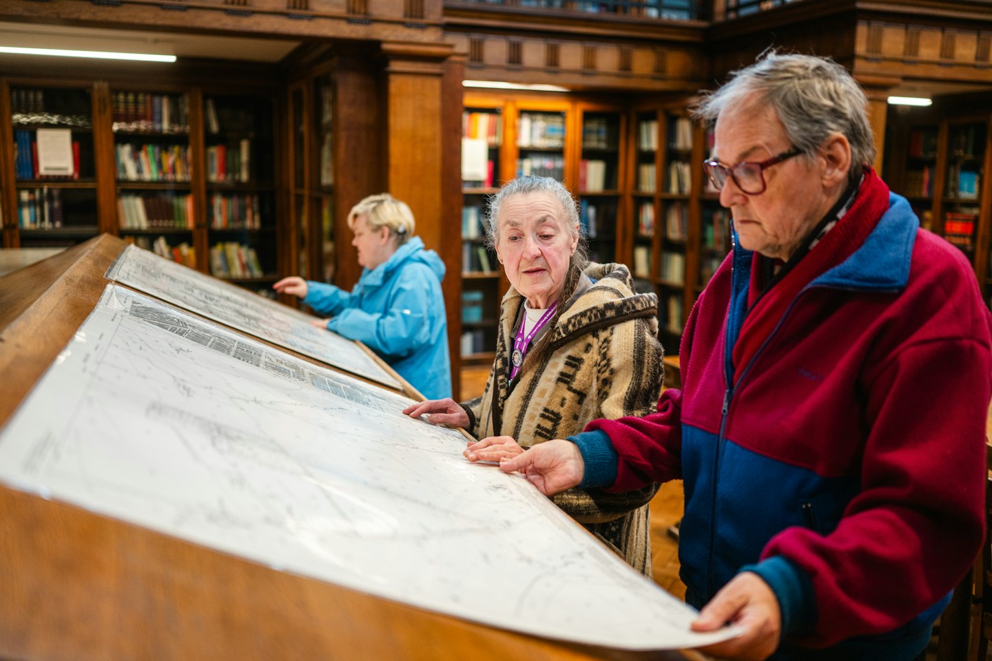 People examine historical maps on wooden tables in a library.